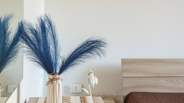 A room with a white wall, a wooden nightstand, a white vase with blue feathers, a brown bed, and a white lamp. - Casa Clale (Marina di Pescoluse)