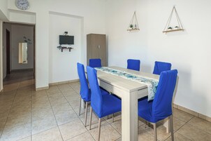 Un comedor con una mesa, sillas azules, un armario de madera, un televisor montado en la pared y una planta colgante pequeña.