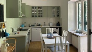 Une cuisine avec des meubles de couleur verte, un plan de travail blanc, un luminaire suspendu, une table à manger avec des chaises et une fenêtre ornée de barreaux en fer décoratifs.