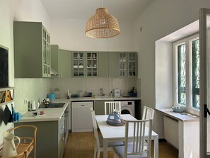 Eine Küche mit grünen Schränken, einer weißen Arbeitsplatte, einer Pendelleuchte, einem Esstisch mit Stühlen und einem Fenster mit schmiedeeisernen Gitterstäben.
