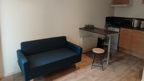 Ett kompakt bostadsutrymme med en soffa, en liten pall, ett litet kök med mikrovågsugn och ett skåp.