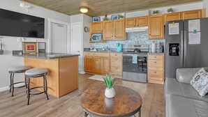 A modern kitchen with wooden cabinets, a stainless steel refrigerator, a microwave, and a large island with stools. - STUNNING! 1 Bedroom Panoramic Ocean View! Beach Cottage Feel, Steps to Shore! (Cocoa Beach)