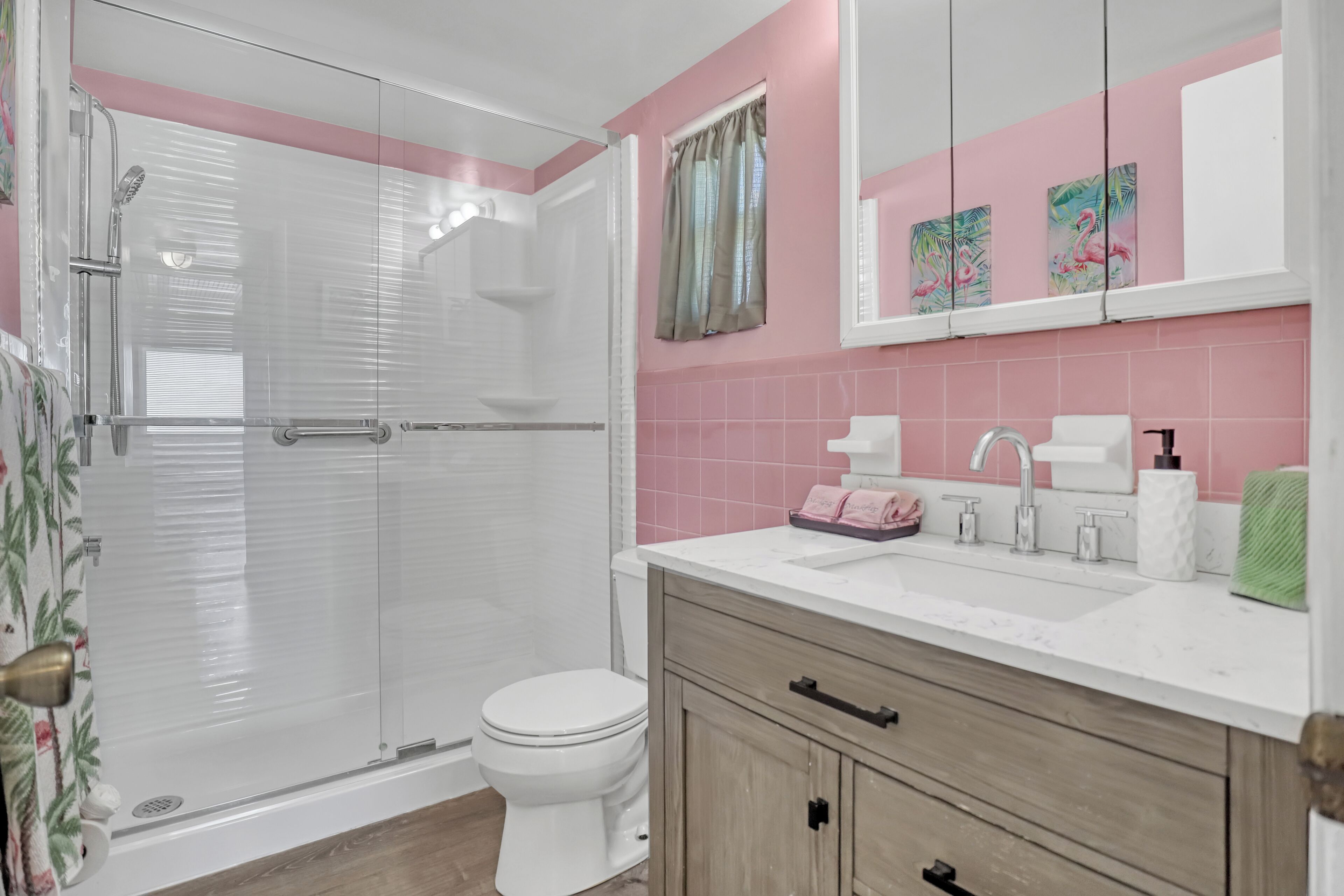 A bathroom with a glass shower enclosure, a toilet, a sink with a mirror above, and pink-tiled walls.