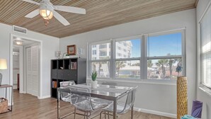 A room with a ceiling fan, a dining table with clear chairs, a bookshelf, and a window with blinds. - STUNNING! 1 Bedroom Panoramic Ocean View! Beach Cottage Feel, Steps to Shore! (Cocoa Beach)