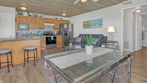 A modern kitchen with wooden cabinets, a central island with stools, a dining table with chairs, a ceiling fan, and a painting on the wall. - STUNNING! 1 Bedroom Panoramic Ocean View! Beach Cottage Feel, Steps to Shore! (Cocoa Beach)