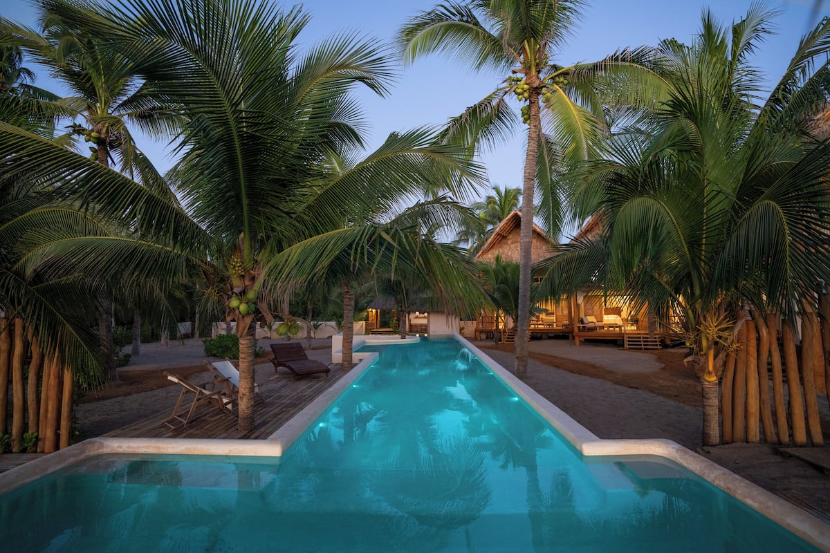 Una piscina con palmeras, una estructura de techo de paja y sillones de descanso.
