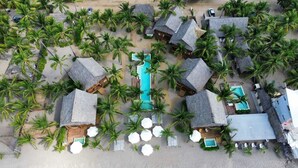 An aerial view of a resort with thatched-roof bungalows, a central swimming pool, and palm trees. - Mangle Rojo Palafitos (Boca de Iguanas)