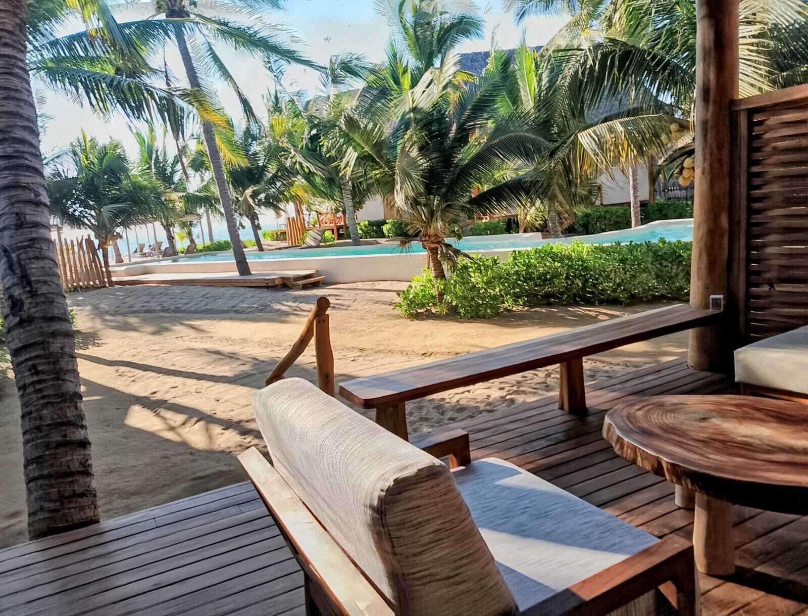 A wooden deck with benches and a round table, overlooking a pool area with palm trees and a sandy beach.