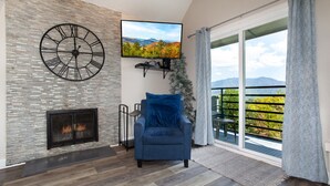 A modern living room with a stone fireplace, a large wall clock, a flat-screen TV, and a balcony with a view of the mountains.