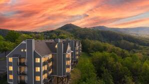 Un hotel de varios pisos con balcones, con vistas a una ladera boscosa y un cielo al atardecer.
