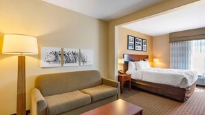 Une chambre d’hôtel avec un canapé, une table en bois, un lit avec une tête de lit, une table de chevet, une lampe et des tableaux encadrés accrochés au mur.
