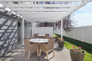Un patio techado con una mesa redonda y sillas de mimbre, plantas en macetas y una valla blanca al fondo.