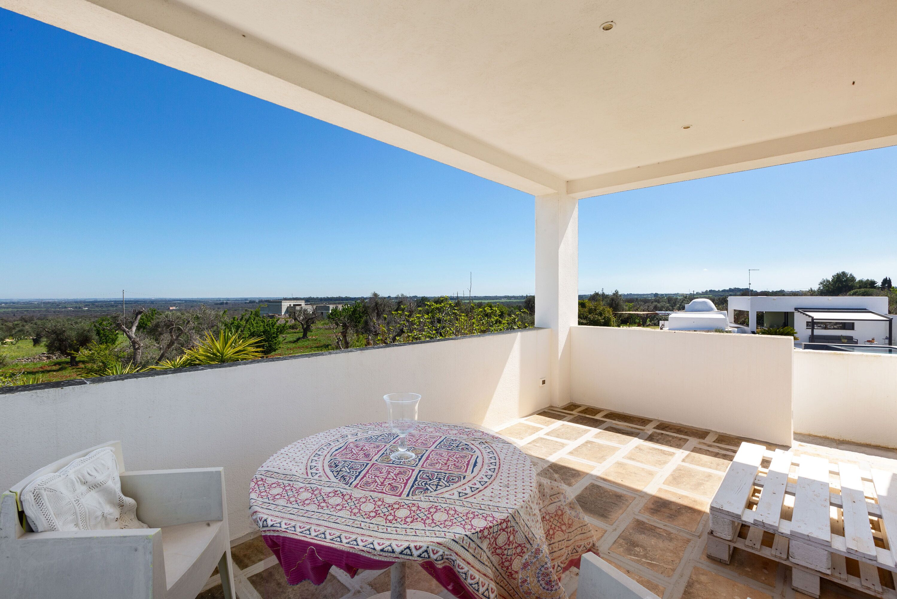 Un balcón con mesa y sillas, vista al campo y un edificio blanco al fondo.