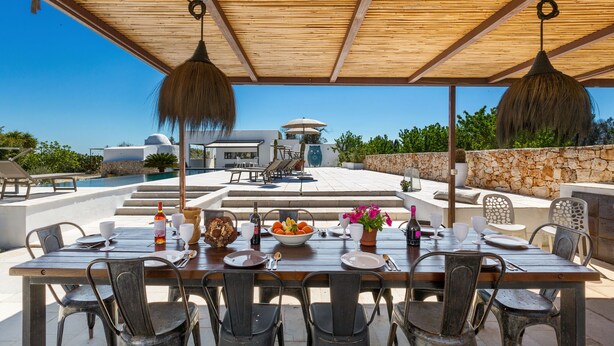 An outdoor dining area with a wooden table set for six, surrounded by chairs, under a wooden pergola with hanging lamps. - Villa Aspri Luxury by Perle di Puglia (Carovigno)