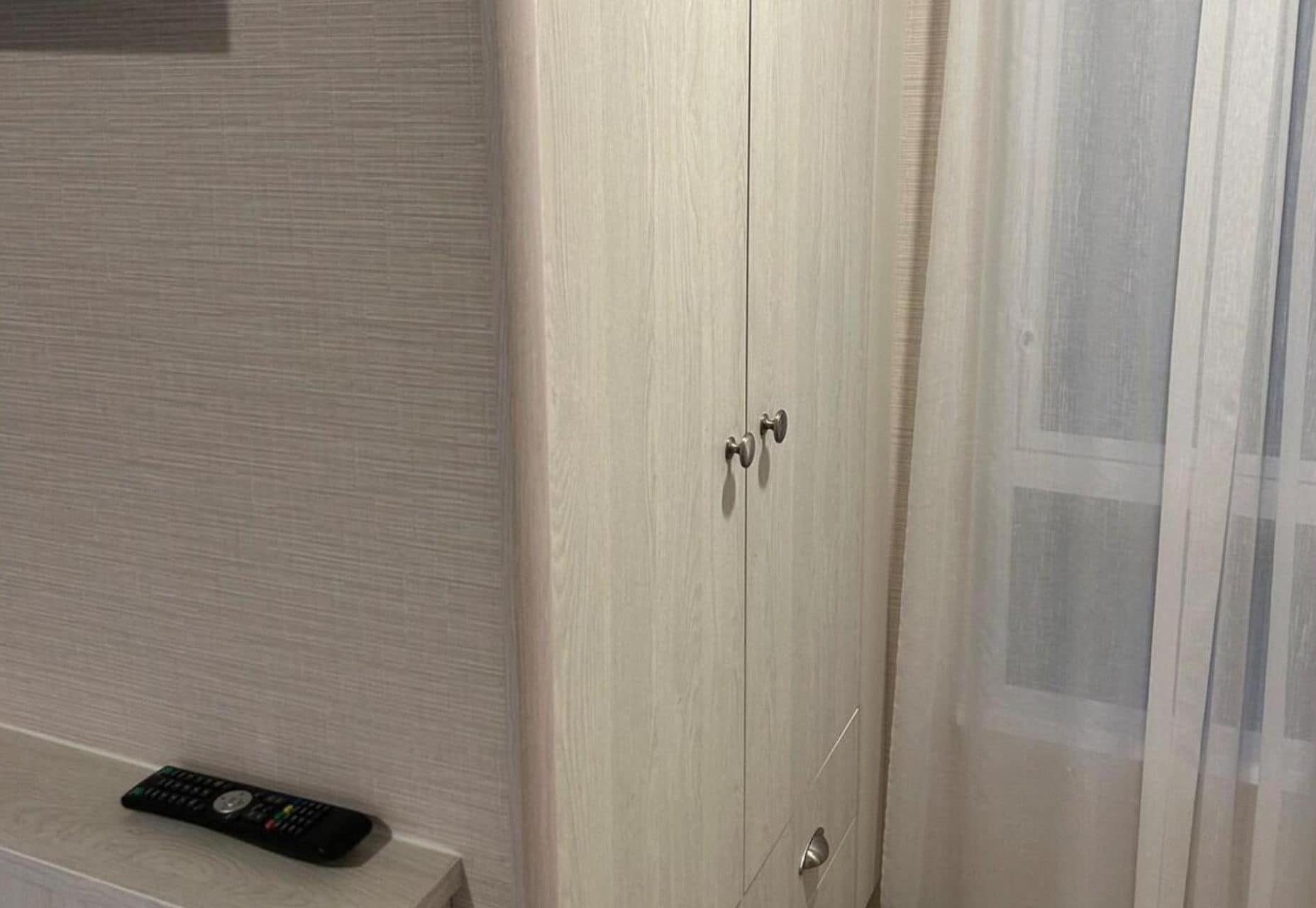 A wooden wardrobe with two doors and a remote control on the floor beside it.