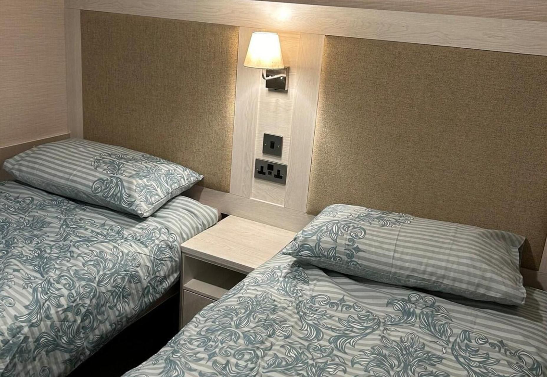 A twin room with two beds, a nightstand, a wall-mounted lamp, and electrical outlets.