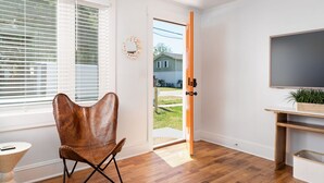 Een kamer met een houten vloer, een bruine lederen stoel, een televisie op een houten plank en een deur die naar buiten leidt.