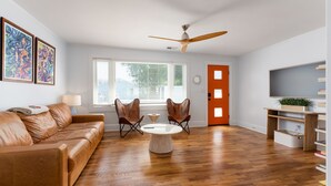 Een moderne woonkamer met een bruine leren bank, twee vleugelstoelen, een ronde salontafel, een plafondventilator, een groot raam met jaloezieën en een boekenkast met een plant.