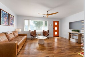Ein modernes Wohnzimmer mit einer braunen Ledersofa, zwei Ohrensesseln, einem runden Couchtisch, einem Deckenventilator, einem großen Fenster mit Jalousien und einem Bücherregal mit einer Pflanze.