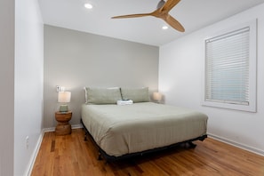 Une chambre à coucher avec un lit, un plancher en bois, un ventilateur de plafond, une lampe de chevet et une fenêtre avec des stores.