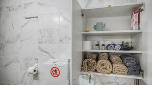 Une salle de bain carrelée de marbre, avec un distributeur de papier toilette, un panneau indiquant qu’il faut se laver les mains sans toucher, et une étagère contenant des serviettes et des articles de toilette.