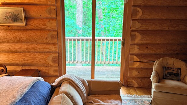 A log cabin room with a sofa, a chair, a bed, and a view of a wooden deck and greenery outside.