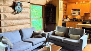 A log cabin interior with two grey sofas, a wooden coffee table, a kitchen area with wooden cabinets, and a large window with a view of greenery.