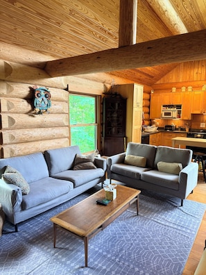 A log cabin interior with two grey sofas, a wooden coffee table, a kitchen area with wooden cabinets, and a large window with a view of greenery.