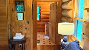 A log cabin interior with a bed, a table with towels, a lamp, and a window with a view.