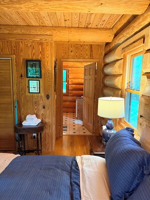 A log cabin interior with a bed, a table with towels, a lamp, and a window with a view.