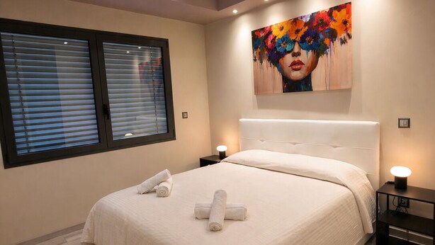 A modern bedroom with a large bed, two bedside tables, a window with blinds, and a colorful painting on the wall.