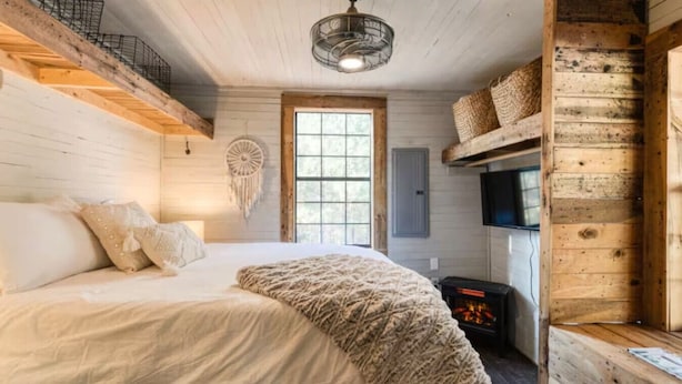 A cozy cabin bedroom with a bed, a wall-mounted TV, a fireplace, and a large window. - Dreamy Mountaintop Escape for Two | Sauna & Views (Blue Ridge)