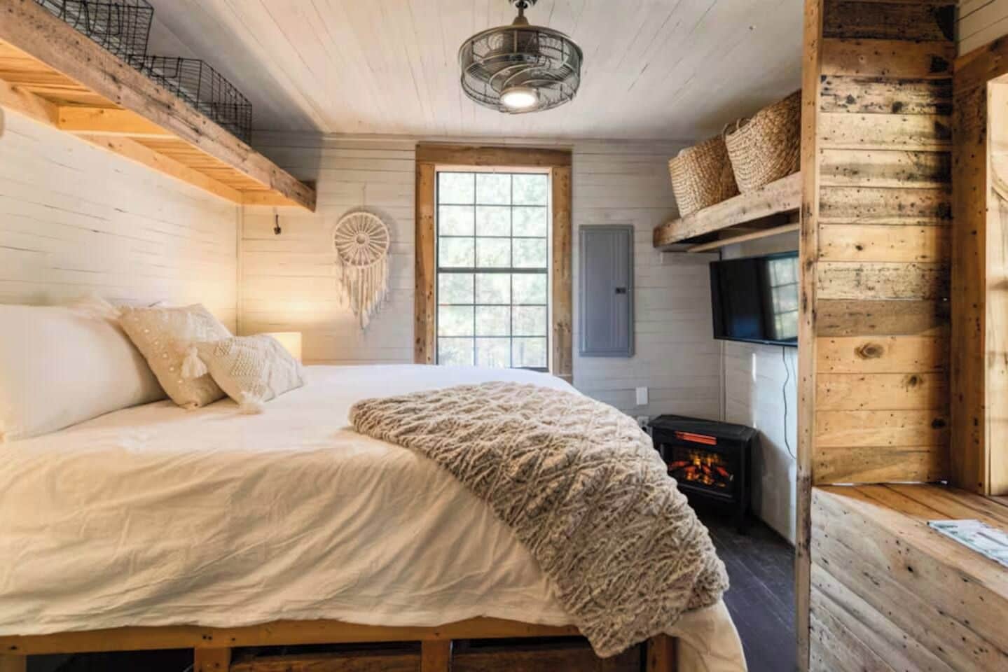 A cozy bedroom with a bed, a wall-mounted TV, a fireplace, and a large window.