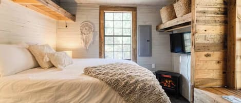 Un dormitorio acogedor con cama, televisor en la pared, chimenea y ventana grande.