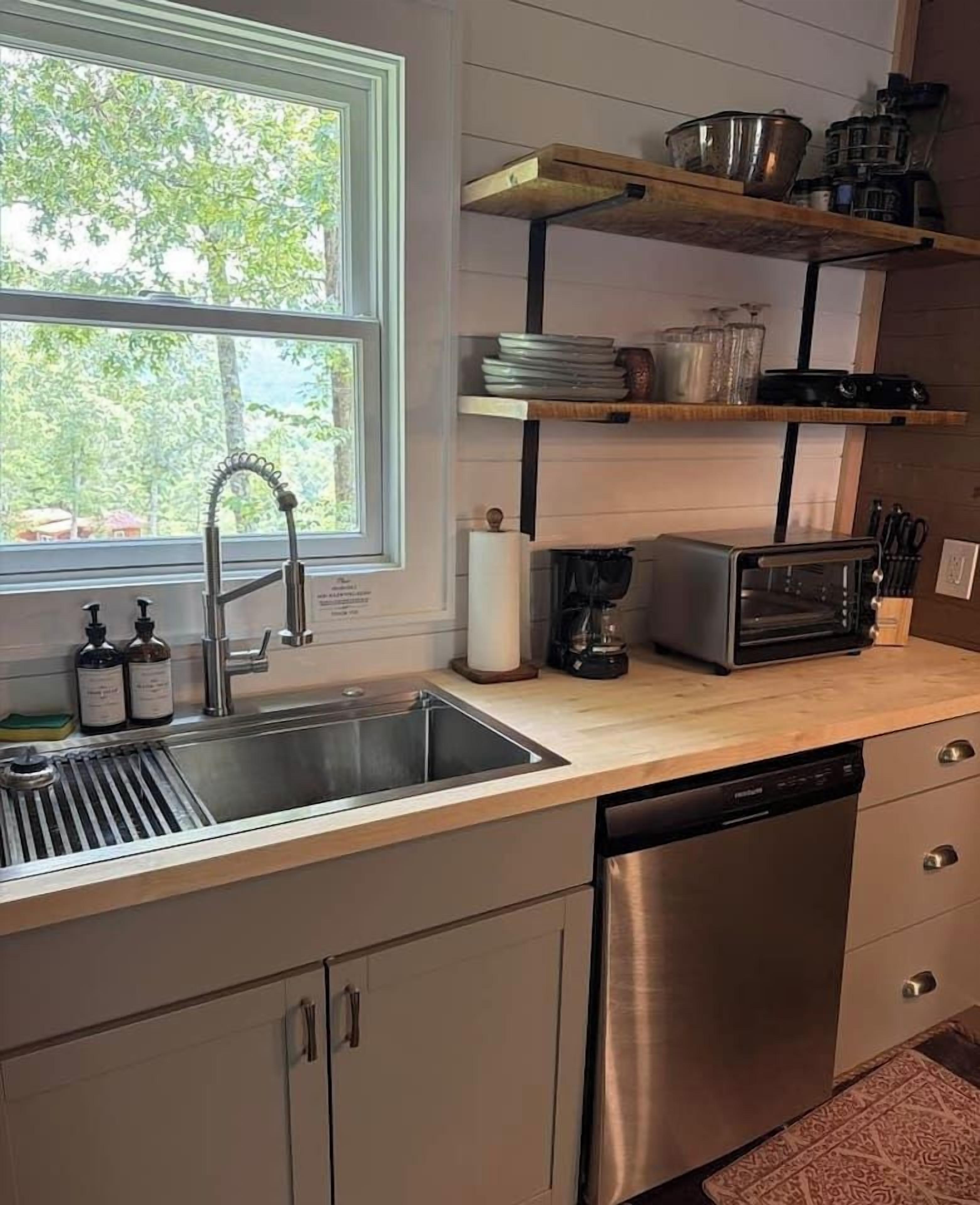 A kitchen with a stainless steel sink, a dishwasher, a coffee maker, and a toaster. There are wooden shelves above the sink and a window with a view of trees.