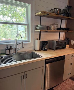 Eine Küche mit einer Spüle aus Edelstahl, einem Geschirrspüler, einer Kaffeemaschine und einem Toaster. Oberhalb der Spüle befinden sich Holzregale und ein Fenster mit Blick auf Bäume.
