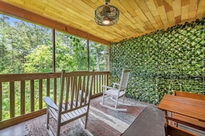 A covered porch with a wooden ceiling, a ceiling fan, a wooden bench, a rocking chair, and a table.
