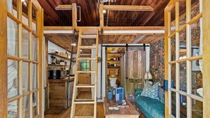 A rustic interior with wooden beams, a lofted sleeping area, a kitchen with shelves, and a cozy seating area with a sofa and table. - Gilded Hideaway | Romantic Tiny Cabin in the Woods (Blue Ridge)