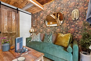 Ein gemütliches Zimmer mit einem grünen Sofa, Holzboden, floralem Wandpapier und einer Holztür.