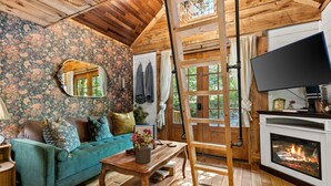A cozy room with a wooden ceiling, floral wallpaper, a teal sofa, a wooden table with a vase, a fireplace, and a TV. - Gilded Hideaway | Romantic Tiny Cabin in the Woods (Blue Ridge)
