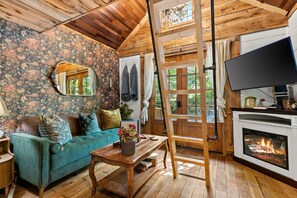 A cozy room with a wooden ceiling, floral wallpaper, a teal sofa, a wooden table with a vase, a fireplace, and a TV.