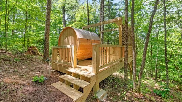 A wooden cabin with a barrel-shaped structure, wooden deck, and steps leading up to it, surrounded by a forest. - Gilded Hideaway | Romantic Tiny Cabin in the Woods (Blue Ridge)