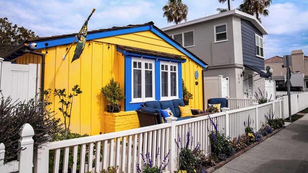 Een geel huis met een blauwe deur en ramen, een wit hekwerk en een veranda met een blauwe stoel.