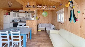 A wooden cabin interior with a dining table, chairs, a sofa, a kitchen area, and a wall decorated with bottles and lights. - 8-BERTH CHALET WITH LARGE TERRACE (Gruissan)