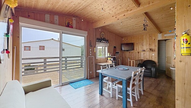 A wooden cabin with a dining area, a sofa, a TV, and a view of a building outside. - 8-BERTH CHALET WITH LARGE TERRACE (Gruissan)