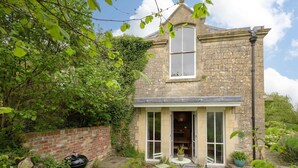 Une maison en pierre avec une grande fenêtre, un mur en briques et un jardin agrémenté d’un barbecue et de chaises.
