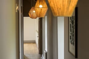 Un couloir se terminant par une porte en bois, un tableau encadré au mur et un luminaire suspendu au plafond.