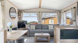 A cozy living area with a sofa, TV, and table, a mirror, and large windows with curtains. - Cosy Lodge in North Devon with Private Parking (Ilfracombe)