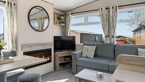 A cozy living area with a fireplace, sofa, TV, and a view of the outside. - Cosy Lodge in North Devon with Private Parking (Ilfracombe)