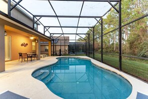 Una piscina moderna al aire libre con cerramiento de cristal, un comedor con sillas y vistas a los árboles circundantes.
